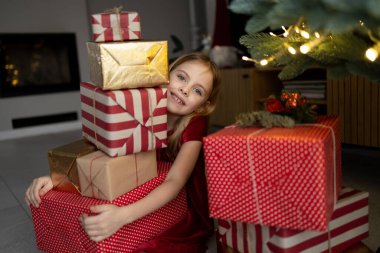 Kırmızı elbiseli küçük bir kız gülümserken bir sürü renkli Noel hediyesini kucaklıyor. Arka planda dekore edilmiş ağaç ve şömine, sıcak ve neşeli tatil ortamı, kopyalama alanı