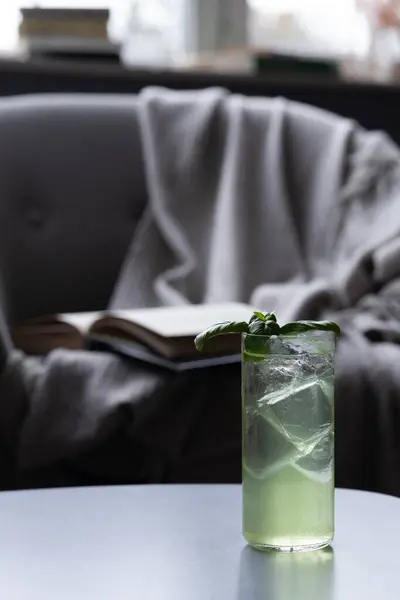 Yumuşak battaniyeyle örtülmüş rahat koltuğun yanındaki masada yeşil kokteyl ile dolu uzun bardak, açık kitap, dinlenmek için huzurlu bir atmosfer yaratıyor.