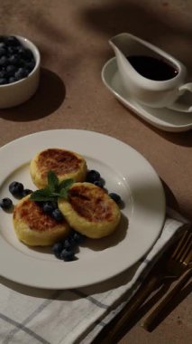 Taze yaban mersini ve nane filizi ile süslenmiş üç tüylü kuark gözlemesi. Masada küçük bir kase şurup var. Arka planda rüzgarda sallanan yaprakların gölgeleri