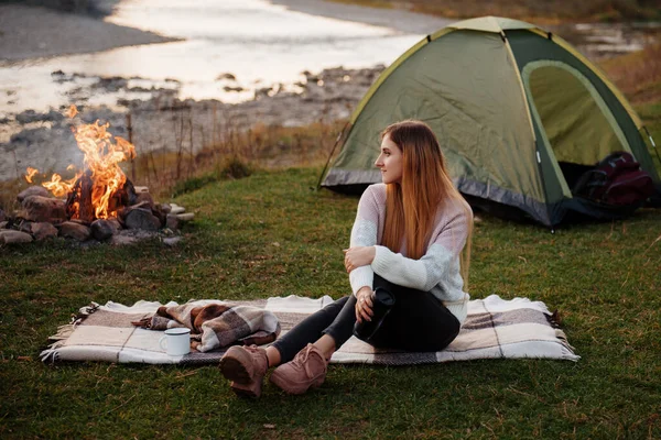 Young woman sits on a blanket on the grass near green tourist tent and bonfire. Girl drinks hot tea or coffee from thermos cup on the background of river and mountains. Resting and hiking concept