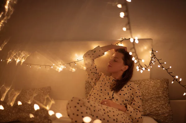 Relaxed woman laying in the bed in darkness with glowing lights. Woman sitting in the bedroom in evening. Calm atmosphere concept