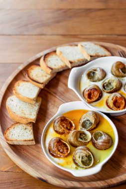 Two plates of baked snails sit on a large brown wooden plate with croutons. Snails baked with butter and spices. Brown background. Exquisite food. French cuisine concept.