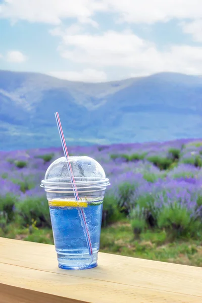 A misted glass with lavender lemonade stands on a beige board against the background of a lavender field, mountains and a cloudy sky