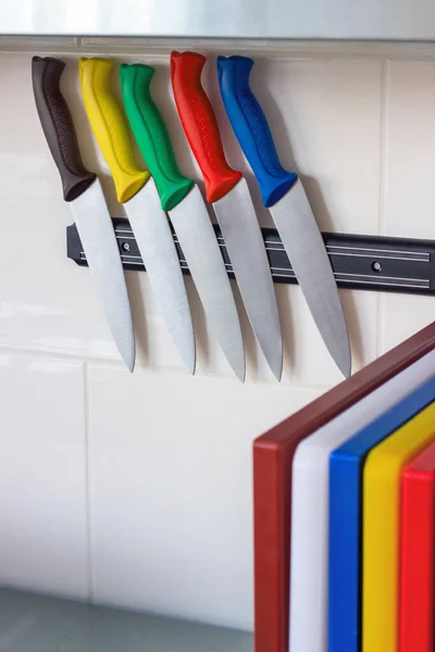 Five kitchen knives with colorful handles hang on a magnetic holder. Multicolored kitchen cutting boards. Professional kitchen equipment for chefs.Food safety. HACCP. Place for text