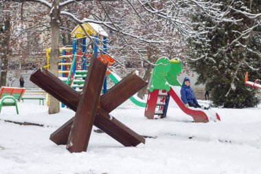 Snow-covered playground in the courtyard of a residential building in Lviv Ukraine. An anti-tank hedgehog is installed nearby. War in Ukraine. A little Ukrainian boy is playing on a slide. 14.12.2022