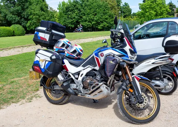 Galzignano Terme, Padua, İtalya-17 Mayıs 2025: Çakıl yolu üzerinde yeşil ağaçlar ve çimlerle çevrili renkli eyer çantaları ve kasklarla donatılmış macera motosikleti Honda doğada heyecan verici bir yolculuğa hazır.