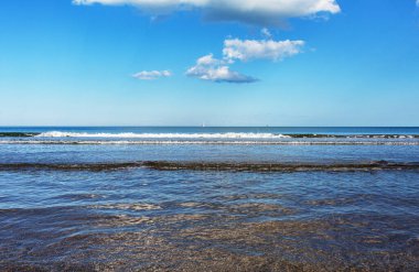 Sahilde hafif dalgaların olduğu sakin bir sahil sahnesi, bulutlu parlak mavi gökyüzü, rahatlamayı davet ediyor ve doğa severler için huzurlu bir ortam.