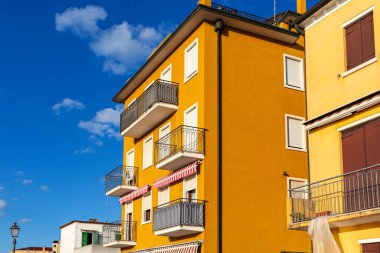 Parlak sarı apartman antik şehir Sottomarina Venedik İtalya 'da balkonları ve çizgili tenteleri açık mavi gökyüzü altında modern mimari ve canlı şehir atmosferini gözler önüne seriyor.