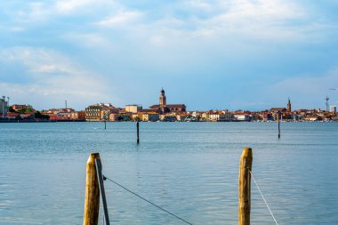  İtalya 'nın sakin bir su fonu olan Chioggia manzarası. Önünde ahşap direkler var. Uzaklarda büyüleyici binalar ve kilise kulesi sergileniyor.