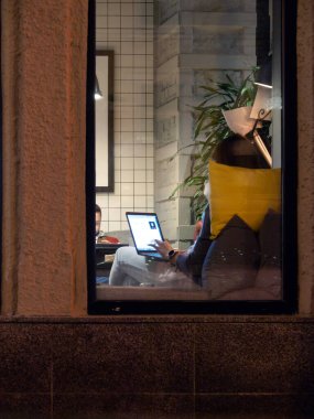 Evening coworking. A girl with a laptop from the back, a moment before pressing a key that interrupts or activates a video session. In the back, a guy with a mobile phone in his hand is barely noticeable.