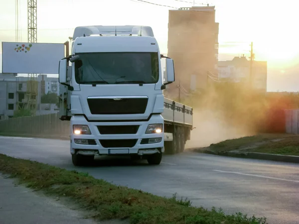 A truck pulls out onto the road. On the left is a house under construction and a blank banner with hearts. There may be your inscription. In the back, dust clouds are visible in the rays of a setting sun.