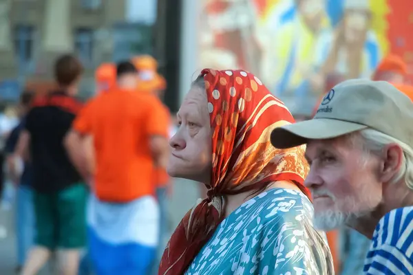 Kharkiv, Ukrayna-13 Haziran 2012: Bir büyükannenin kafasında başörtüsü, bir dedesinin de beyzbol şapkası vardır. Yaşlı bir erkek ve kadın, Euro 2012 boyunca ulusal bayrağı taşıyan bir Hollandalı taraftarın arka planına karşıdır.