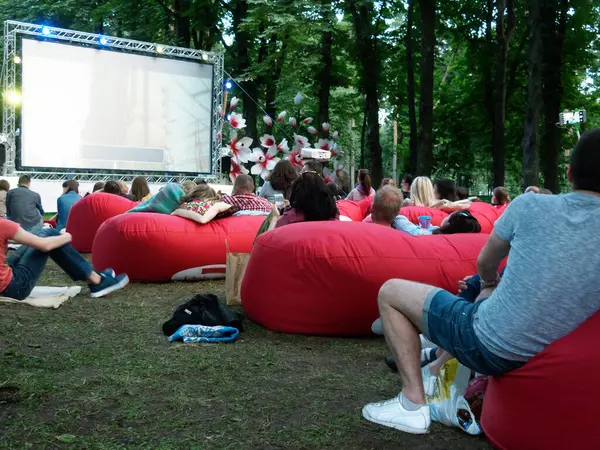 Açık hava yaz sineması. Bir grup genç, dışarıda, parktaki yumuşak kırmızı yastıkların üzerinde bir çeşit video dizisi gösteren dev bir ekranın yanında oturuyorlar. Ekranın altında yazıtın için boş bir gri alan var. 