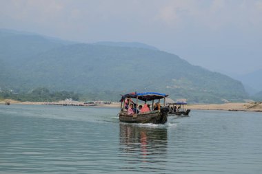 İki geleneksel tekne, uzak dağlar ve canlı yeşilliklerle çerçevelenmiş sakin bir nehirde süzülerek ilerler. Bulutlar öğleden sonra gökyüzünü yumuşakça kaplar..