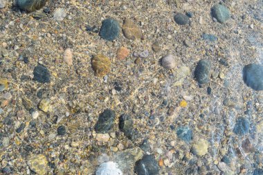 Bir gölün sakin yüzeyi, berrak suyun altında çeşitli renkli çakıl taşlarını sergiliyor. Güzel bir günde güneş ışığını yansıtıyor..