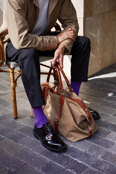 Bir adam dışarıda bir kafede oturuyor ve elinde bir çanta tutuyor ve menekşe çoraplar giyiyor.