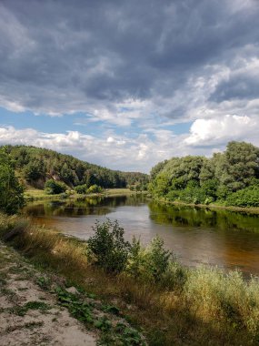 Sakin suları, yeşil ağaçları ve bulutlu yaz gökyüzünü yansıtan huzurlu orman nehri. Yumuşak ışık ve doğal tonlar sakin bir atmosfer yaratır. Doğa, seyahat, rahatlama ve açık hava temalı tasarımlar için ideal.
