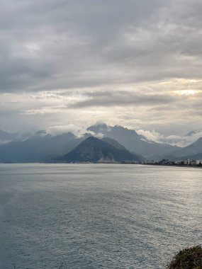 Akdeniz boyunca bulutlarla kaplı dağlarla kaplanmış kasvetli kıyı manzarası. Sakin su ve şehir kıyıları dramatik bir gökyüzü altında. Türkiye 'nin Antalya kentinde deniz manzarası.