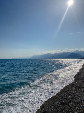Turkuaz suyu ve çakıl taşı plajı boyunca hafif dalgalarla güneşli deniz manzarası. Akdeniz 'e yansıyan güneş ışığı ufukta dağlarla birlikte. Türkiye 'de manzaralı kıyı manzarası.