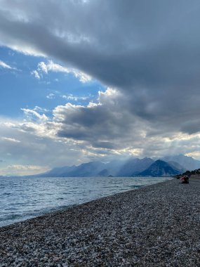 Yoğun bulutların altında çakıl taşı plajı ve dağ sırası olan dramatik Akdeniz kıyıları. Türkiye 'nin Antalya kenti yakınlarında denizden yükselen güneş ışınları. Doğal ışık ve kopyalama alanı olan atmosferik kıyı manzarası.
