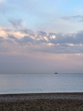 Gün batımında sakin deniz, gökyüzü ve ufukta dramatik bulutlar. Sessiz bir çakıl taşı plajının üzerinde küçük bir tekne silueti. Yumuşak akşam ışığı ve sakin atmosferi olan asgari deniz manzarası.