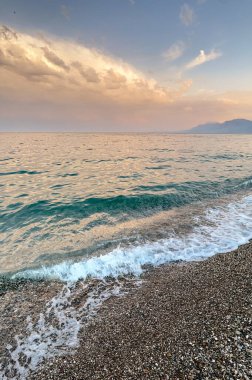 Türkiye 'de günbatımında Akdeniz' in turkuaz dalgaları çakıl taşı plajını yıkıyor. Uzak dağların üzerinde yumuşak altın ışık ve pastel bulutlar seyahat ve tatil havasıyla huzurlu bir deniz manzarası yaratır..