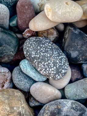 Sahildeki renkli çakıl taşlarının detaylı görüntüsü çeşitli doğal dokuları, şekilleri ve renkleri gösteriyor. Yumuşak kayaların barışçıl düzenlenmesi, doğa, seyahat ve rahatlama temaları için ideal sakin ve organik bir zemin oluşturur..