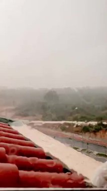 Yağmurun yağışının karamsar bir videosu, ıslak bir pencereden görülüyor. Türkiye 'nin Antalya kentindeki kırmızı kiremitli çatı ve yeşil manzarayı yağmur damlaları bulanıklaştırıyor. Atmosferik klip duygusal, hava durumu veya mevsimsel projeler için mükemmel.