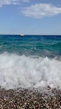 Güçlü deniz dalgaları, parlak mavi bir gökyüzünün altında ufukta görünen sarı bir tekneyle çakıl taşlı bir plaja çarpar. Temiz su, hareket ve doğal ışık seyahat ve doğa projeleri için mükemmel bir kıyı manzarası yaratır..