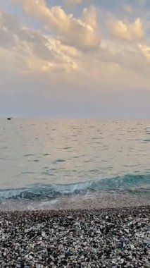 Sakin deniz dalgaları yumuşak bulutlu altın bir gökyüzünün altındaki çakıl taşı plajına yavaşça yuvarlanıyor. Ufukta küçük bir tekne görünüyor. Huzurlu ve manzaralı doğal manzara, seyahat, rahatlama, meditasyon ve arka plan görselleri için ideal.