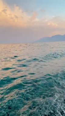 Sakin deniz yüzeyi, yumuşak altın ışıkla pastel bir günbatımı gökyüzünün altında hafif dalgaları yansıtıyor. Uzak dağlar ve dingin renkler seyahat ve rahatlama temaları için mükemmel huzurlu bir kıyı manzarası yaratır..