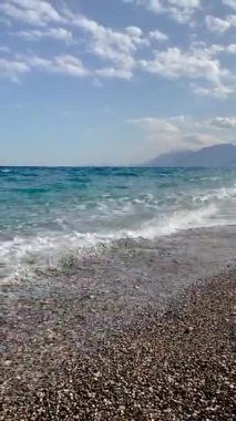 Dalgalar bir çakıl taşı plajına çarpıyor. Uzakta turkuaz deniz ve dağlar var. Dinamik ve manzaralı kıyı manzarası, seyahat, yaz, özgürlük, doğa ve rahatlama temaları için mükemmel..