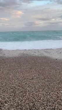 Gün batımında yumuşak bulutlu bir gökyüzünün altında çakıl taşlı kıyıları yıkayan yumuşak turkuaz deniz dalgaları. Huzurlu doğal deniz manzarası, meditasyon, seyahat ve arka plan kullanımı için ideal.