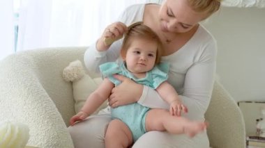Anne, bebeğinin saçlarını tahta saç fırçasıyla tarıyor. Kız ve anne birlikte eğleniyor, gülümsüyor. Çocuk bakımı, ev hayatı rutin, sağlıklı yaşam tarzı