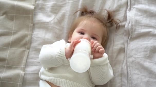 自宅でボトルからミルクを吸う小さな赤ちゃんの女の子 肖像画の幼児の食事 粉ミルク幼児の式を飲む かわいい子供がベッドの上に横たわって ニットセーター ピグテールを着て カメラを見て — ストック動画