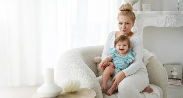 Happy Mother and baby daughter sit in chair in bedroom home. Family look at camera, kid and mummy enjoying time together, smiling. Child care, domestic life routine, healthy lifestyle. Copy space, mock up, template