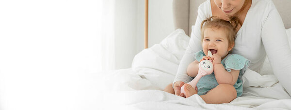 Laughing Mom and baby girl are lying in bed, enjoying time together. Family time concept. Funny playing with toy. Mock up, copy space. Positive babyhood and parenthood.