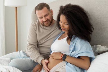 Beyaz adam hamile siyah karısının karnına dokunuyor. Hamile kadın ve sevgi dolu koca evde karnına sarılıyor. Çok ırklı bir çiftin hamile karnındaki bebek hareketini hissediyorum. Yakın plan..