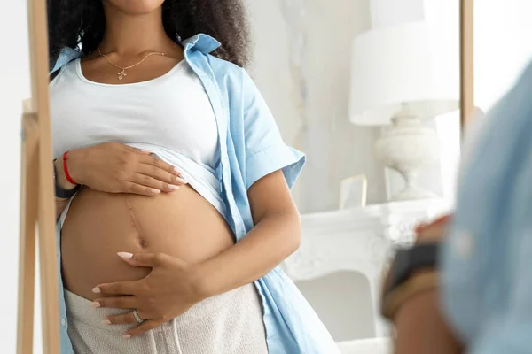 Close up of happy Afro-American pregnant woman with beautiful belly near window at home. Pregnancy, motherhood, future parent, childbirth and expectation concept - pregnancy, motherhood, people and expectation concept
