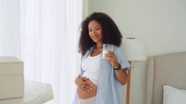 妊娠中の若い女性は窓の近くのカジュアルな服で自宅で牛乳を飲んでいます 妊娠を楽しみ 妊娠中の健康的な食事の概念 乳業栄養の利点 — ストック動画