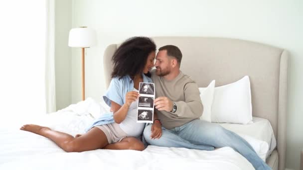 自宅のベッドで超音波画像を見て 多民族妊娠中のカップル 幸せを感じ おなかを撫でて 赤ちゃんの超音波を見て興奮 出産予定日 — ストック動画
