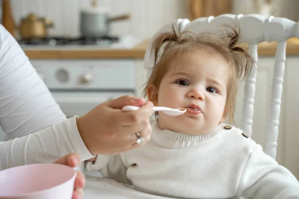 Baby girl eating blend mashed food sitting, on high chair, mother feeding child, hand with spoon for vegetable lunch, baby weaning, first solid food for kid. Healthy eating for baby humans
