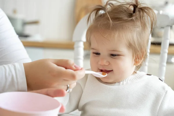 Baby girl eating blend mashed food sitting, on high chair, mother feeding child, hand with spoon for vegetable lunch, baby weaning, first solid food for kid. Healthy eating for baby humans