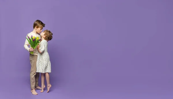Friendly kiss between cute boy and girl standing over purple background, hold spring tulips. Copy space banner for advertisement.