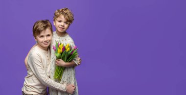 Çocuklar ve kızlar arasında dostça kucaklaşmalar. Mor arka planda duruyorlar. Bahar lalelerini tutuyorlar ve kameraya bakıyorlar. Reklam için boşluk afişini kopyala.