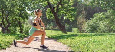 Caucasian middle-aged woman doing workout exercise in public park in sunny summer morning. Warm up before running jogging training. Active healthy lifestyle. Copy space banner