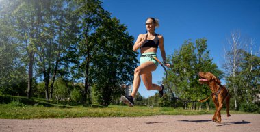 Sporty happy woman jogging with dog outdoor in public park summer. Canicross competition. Healthy life with active pet. Copy space for design