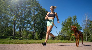 Sporty happy woman jogging with dog outdoor in public park summer. Canicross competition. Healthy life with active pet. Copy space for design