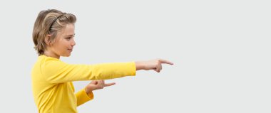 Impressed Emotional Teenager boy happily Joy Showing To Right Over White Background. Copy Space For Advertisement. Excited Blond Kid With Long Hair And Yellow T-Shirt