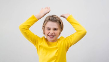 Cheerful Emotional School Boy happily Waving hands up Over White Background. Blond Kid With Long Hair And Yellow T-Shirt Waving Hands Excited. Copy space